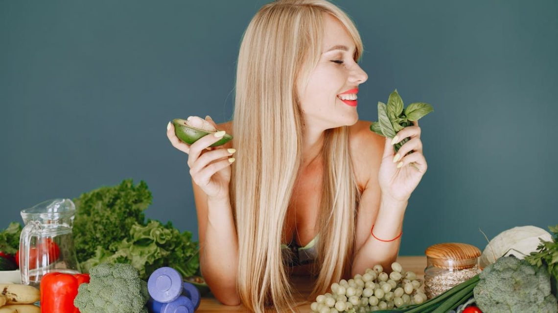 A smiling woman holding avocado and basil, surrounded by fresh vegetables and fruits for healthy eating.