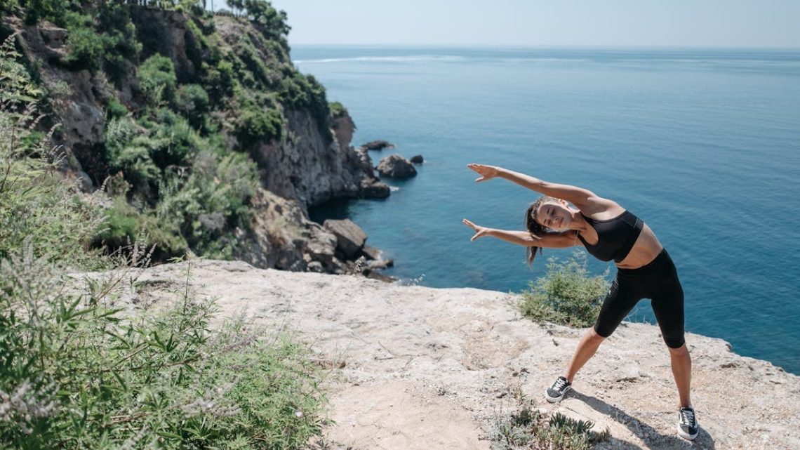 A woman in activewear stretches on a cliff by the ocean, embracing coastal fitness.